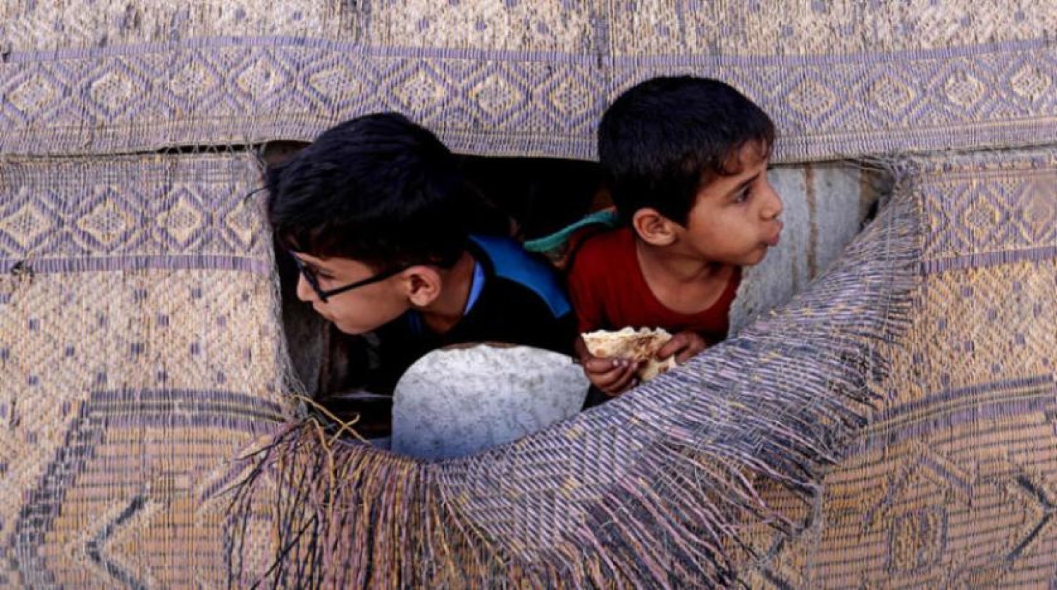 Şati Mülteci Kampı’ndaki çocuklar, yırtık bir hasırdan dışarıya bakıyor (AFP)
